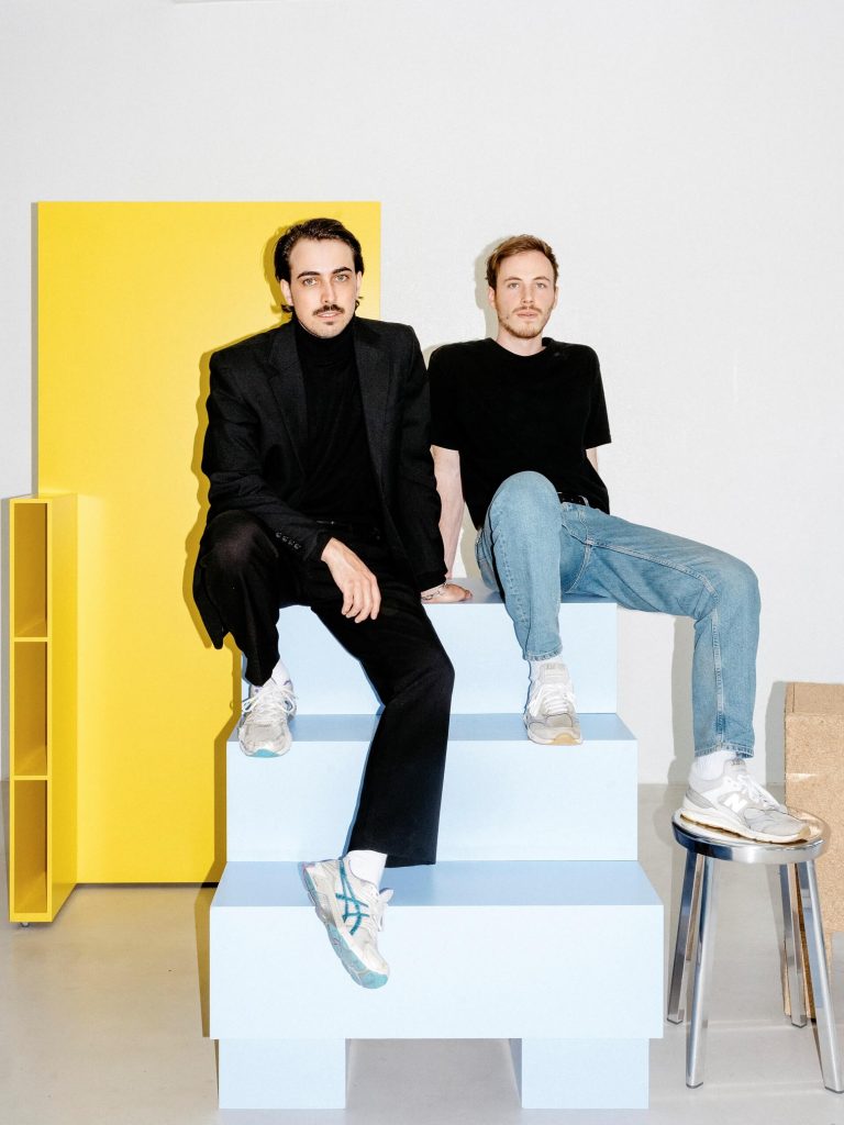 Ganzkörperportrait von Haus Otto (Patrick Henry Nagel und Nils Körner). Die beiden sitzen auf einem von ihnen gestalteten Möbelstück.